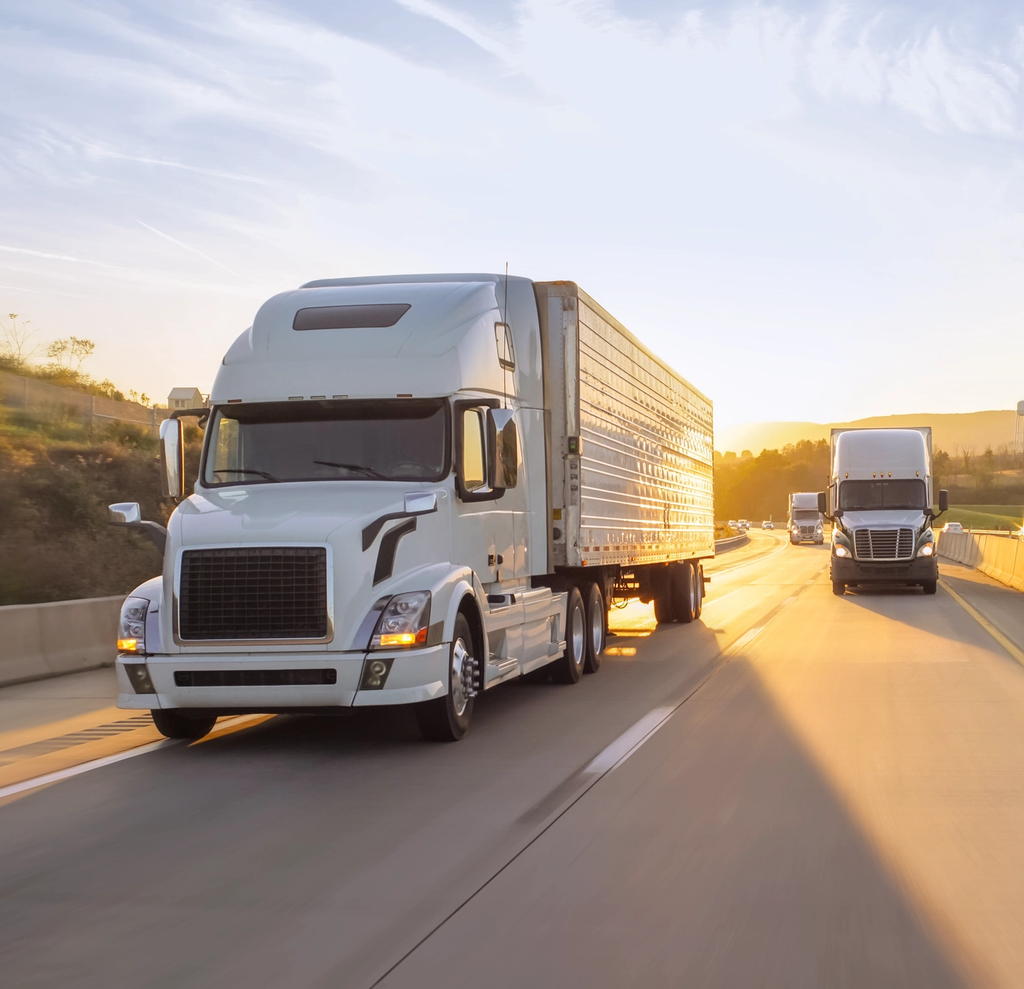 White semi trucks hauling trailers down a highway into a low sunset