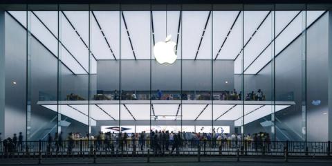 Crowds gather at night outside a glass Apple Store facade lit by a glowing white Apple logo