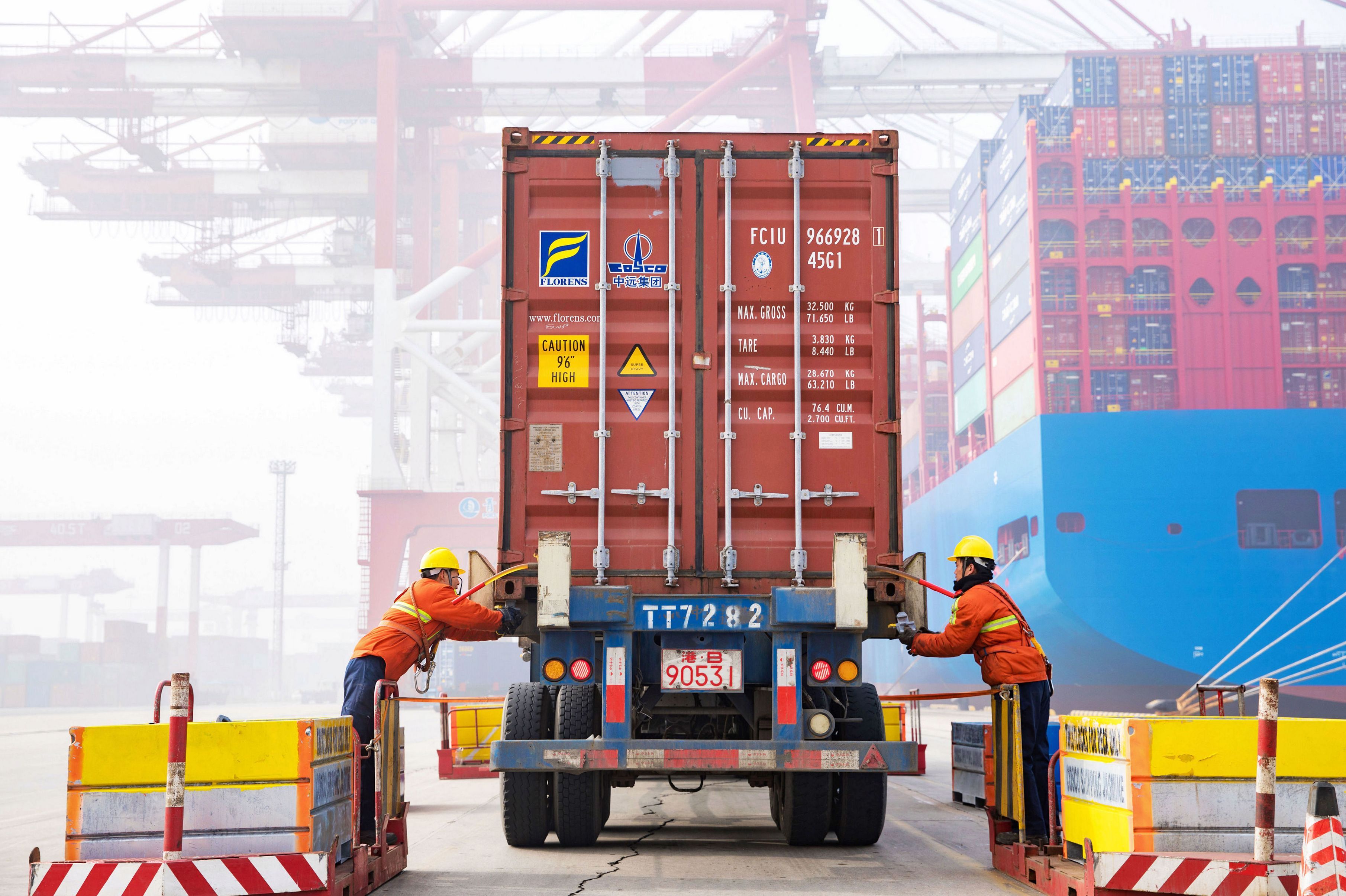 Workers guide a shipping container onto a truck at a container port facility.