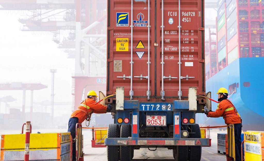 Workers guide a shipping container onto a truck at a container port facility.