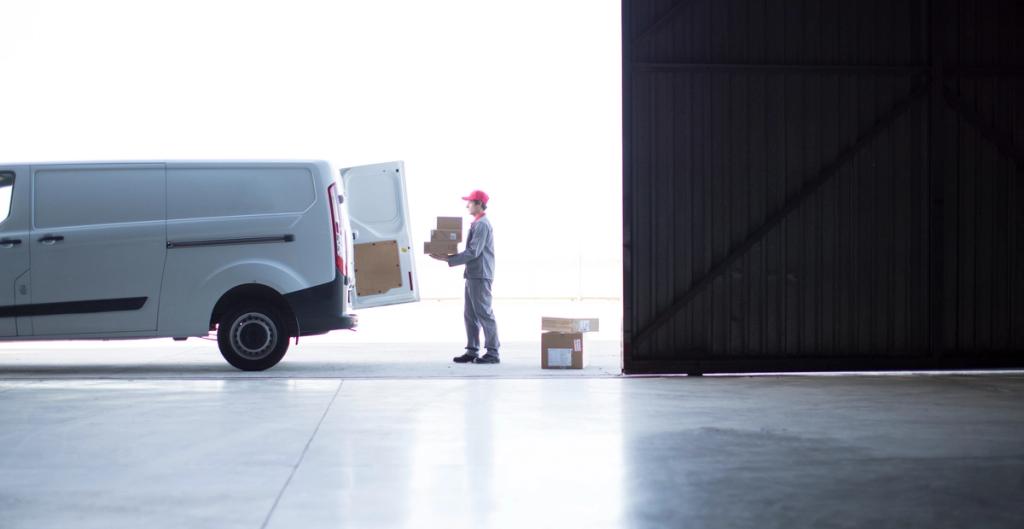 Courier in red cap loads parcels from an open white delivery van into a dark warehouse doorway.