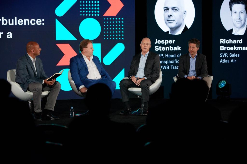Four men on a conference stage panel; screen shows speakers Jesper Stenbak and Richard Broekman with titles.