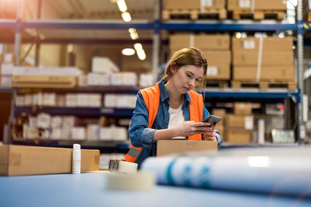 Woman in an orange safety vest checking her phone while leaning on a box in a warehouse aisle