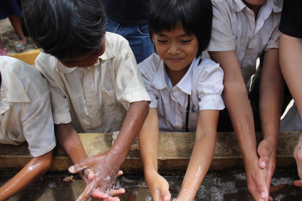 Schoolchildren in uniforms lean over a trough washing soapy hands together outdoors