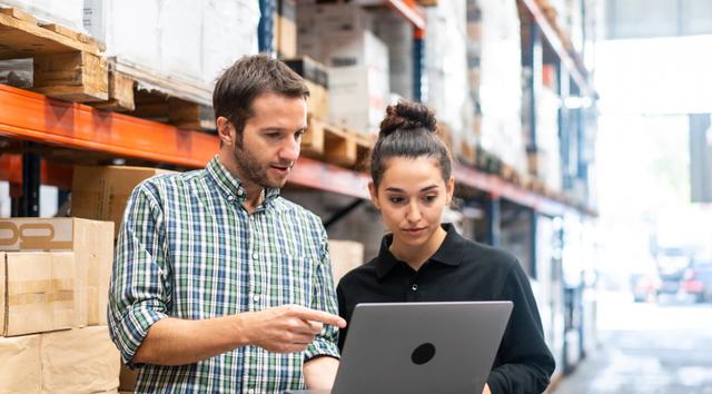 A man and woman reviewing a laptop together in a warehouse storage aisle