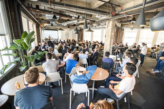 Employees seated at round tables in an open-plan office watching a speaker present beside a projection screen