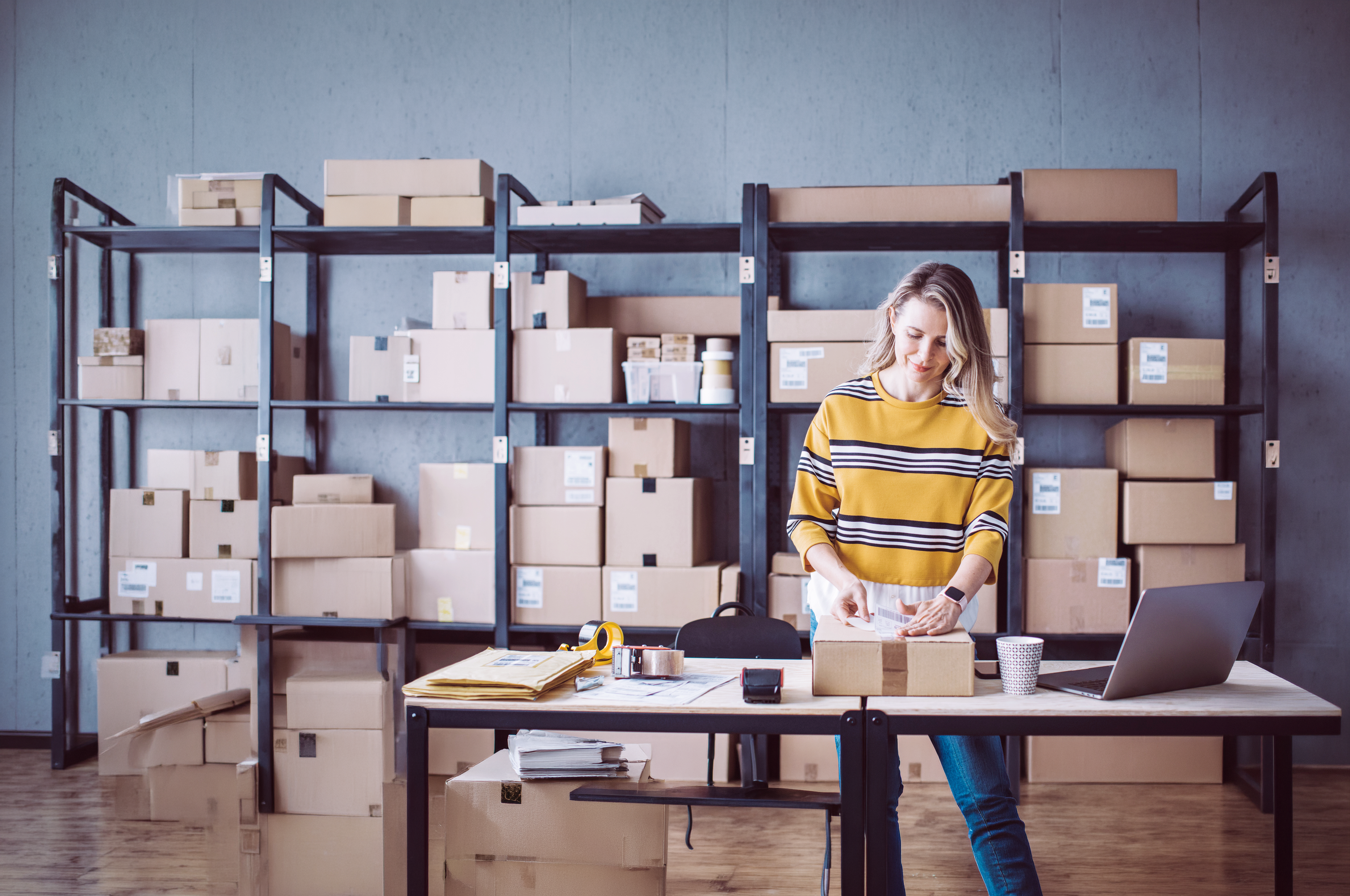 Woman in striped shirt organizing packages at desk with shelving unit full of cardboard boxes behind her.