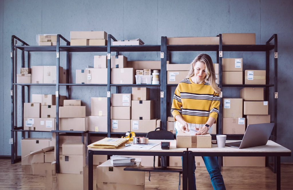 Woman in striped shirt organizing packages at desk with shelving unit full of cardboard boxes behind her.