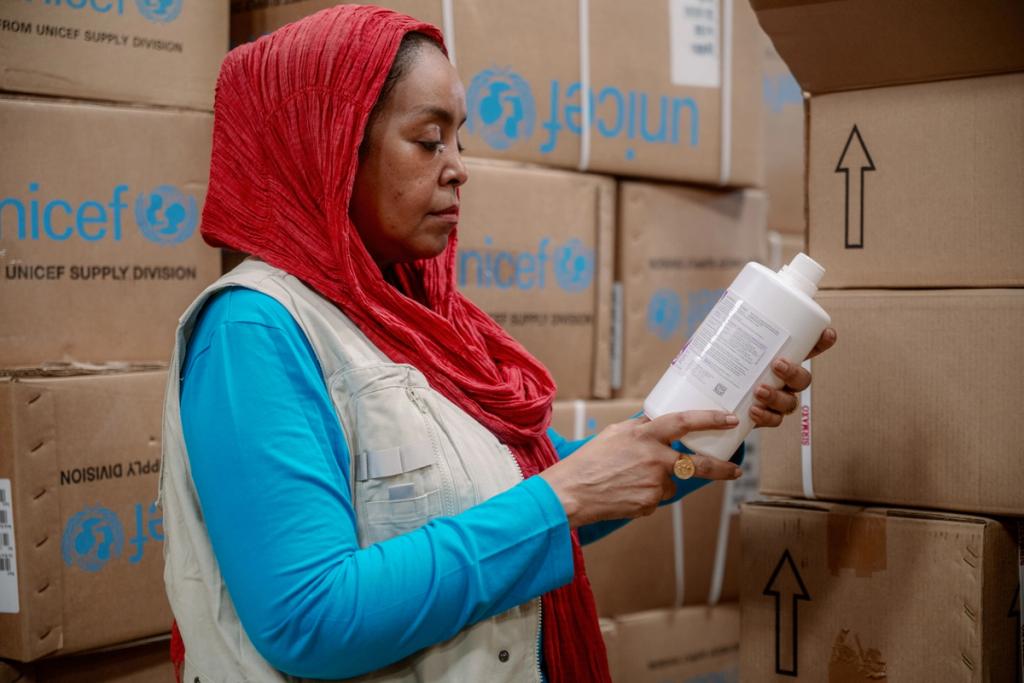 Woman in a red headscarf and aid vest reads a white bottle among stacked UNICEF supply boxes
