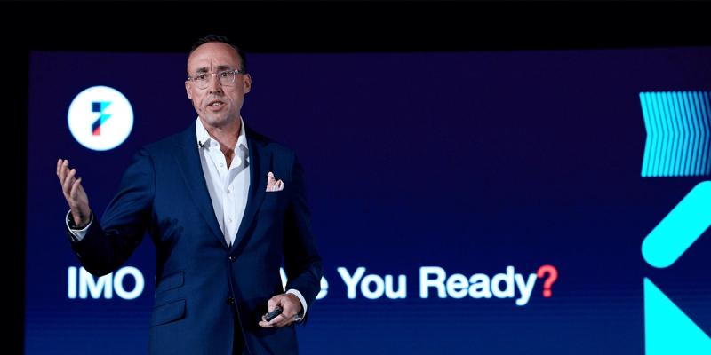 Man in navy suit speaking on stage in front of a blue screen reading IMO Are You Ready with Flexport logo