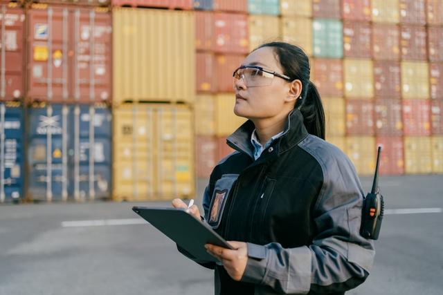 Logistics worker in safety glasses and jacket holding a tablet, looking up at stacked shipping containers at a port yard