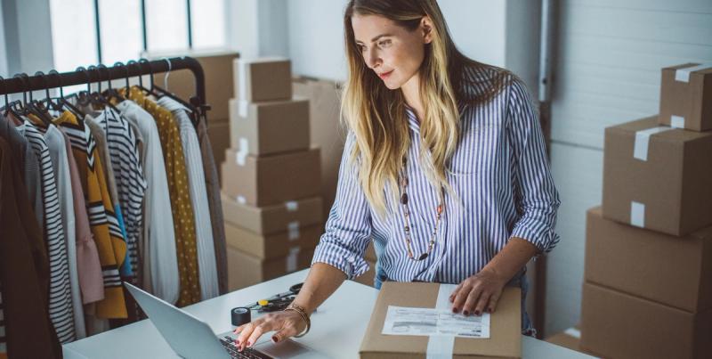 Woman in striped shirt uses a laptop beside a packed shipping box, clothing rack and stacked parcels behind her
