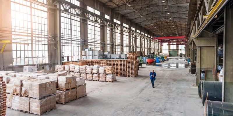 Industrial warehouse interior with a man in a suit walking among stacked pallets of goods under tall windows