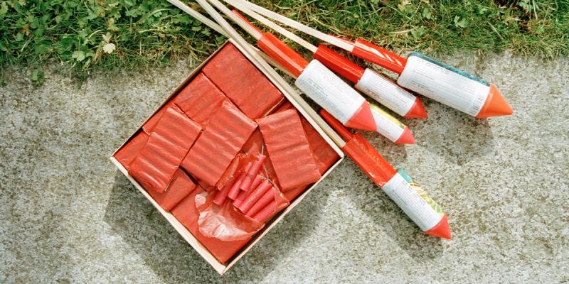 Box of red firecrackers beside several rocket fireworks resting on concrete next to grass