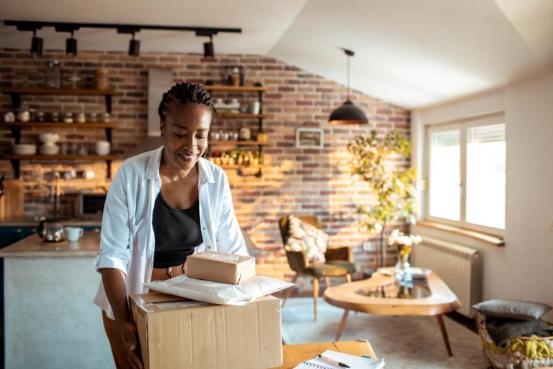 Smiling woman carrying a cardboard box topped with parcels in a sunlit brick-walled home