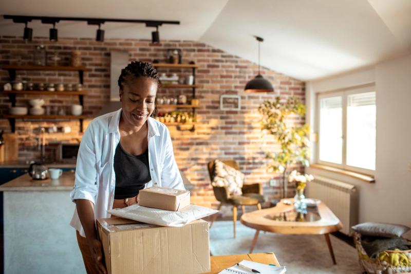 Smiling woman carrying a cardboard box topped with parcels in a sunlit brick-walled home