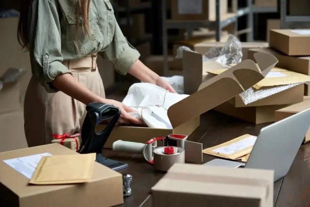 Woman in olive shirt packs an item into a box with a barcode scanner amid parcels and a laptop.