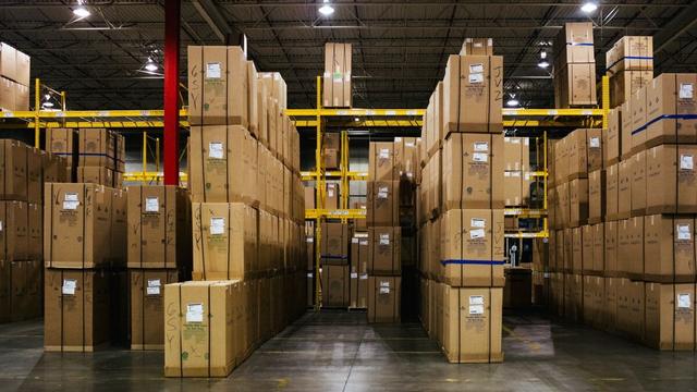 Warehouse interior with tall stacks of cardboard boxes on yellow pallet racking, lit by overhead lights