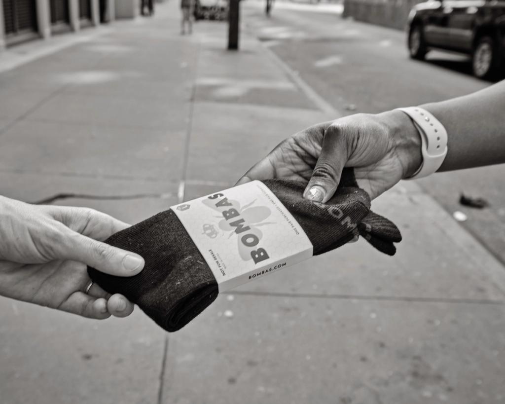 Black-and-white photo of one hand passing a pack of Bombas socks to another on a sidewalk