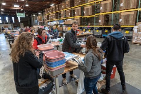 Volunteers sort donated supplies into colored bins at a long table in a warehouse