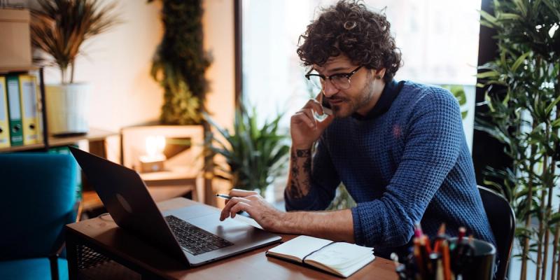 Man in blue sweater and glasses works on a laptop while talking on the phone in a plant-filled office