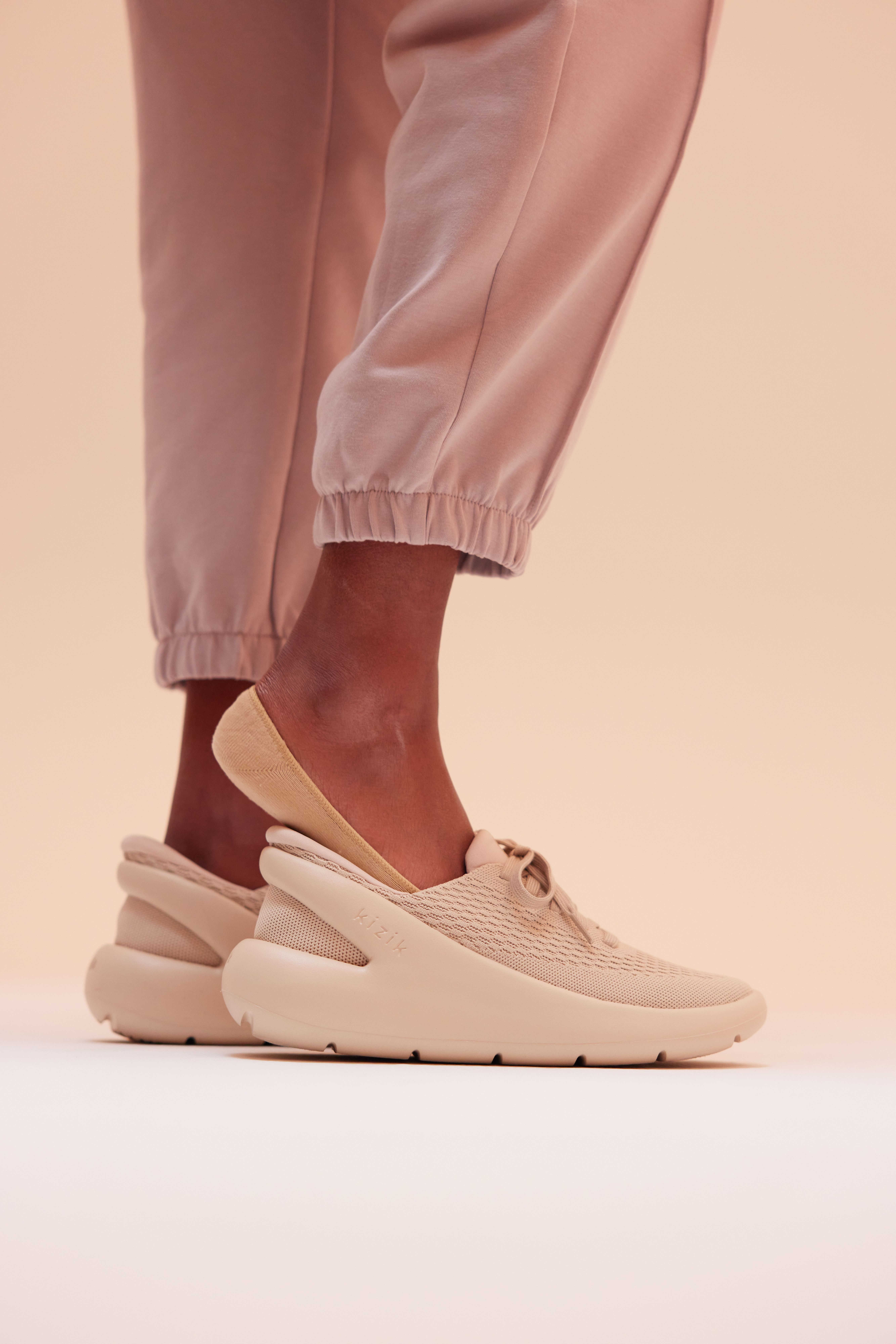Person in pink joggers wears a tan Kizik slip-on sneaker against a neutral backdrop
