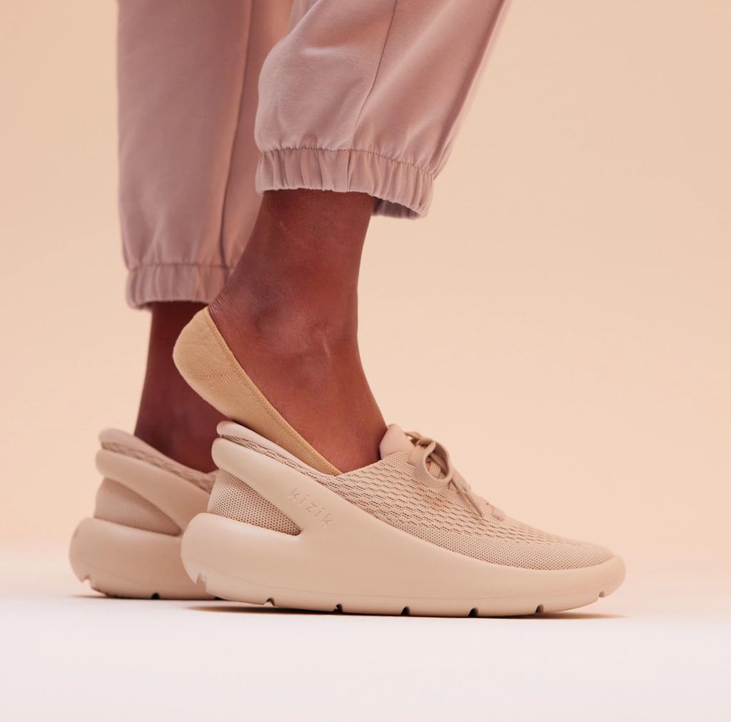 Person in pink joggers wears a tan Kizik slip-on sneaker against a neutral backdrop