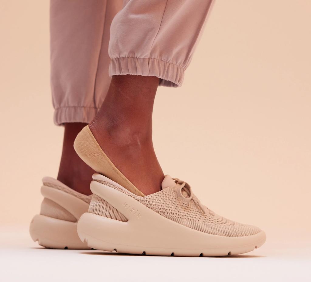 Person in pink joggers wears a tan Kizik slip-on sneaker against a neutral backdrop