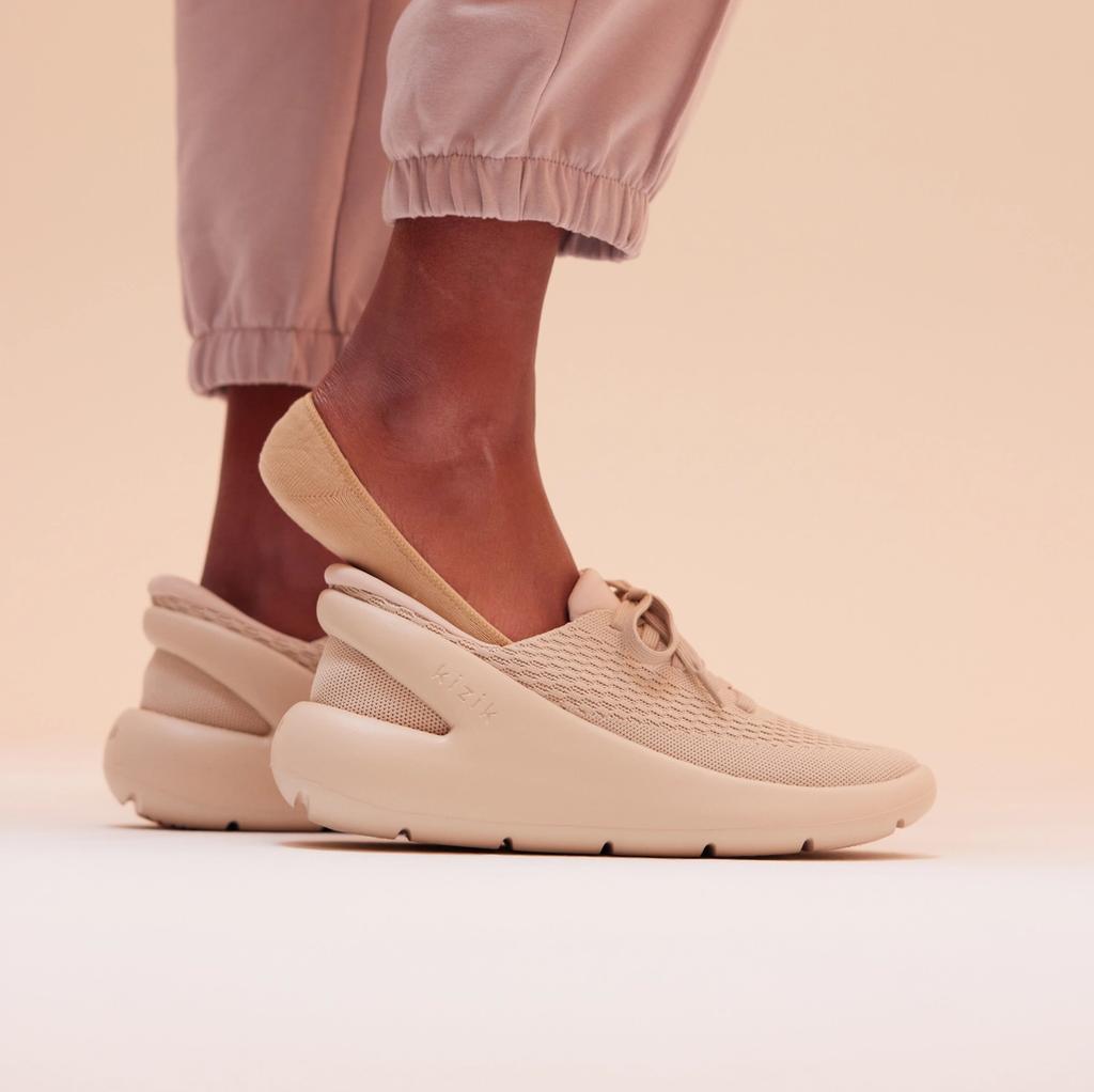 Person in pink joggers wears a tan Kizik slip-on sneaker against a neutral backdrop