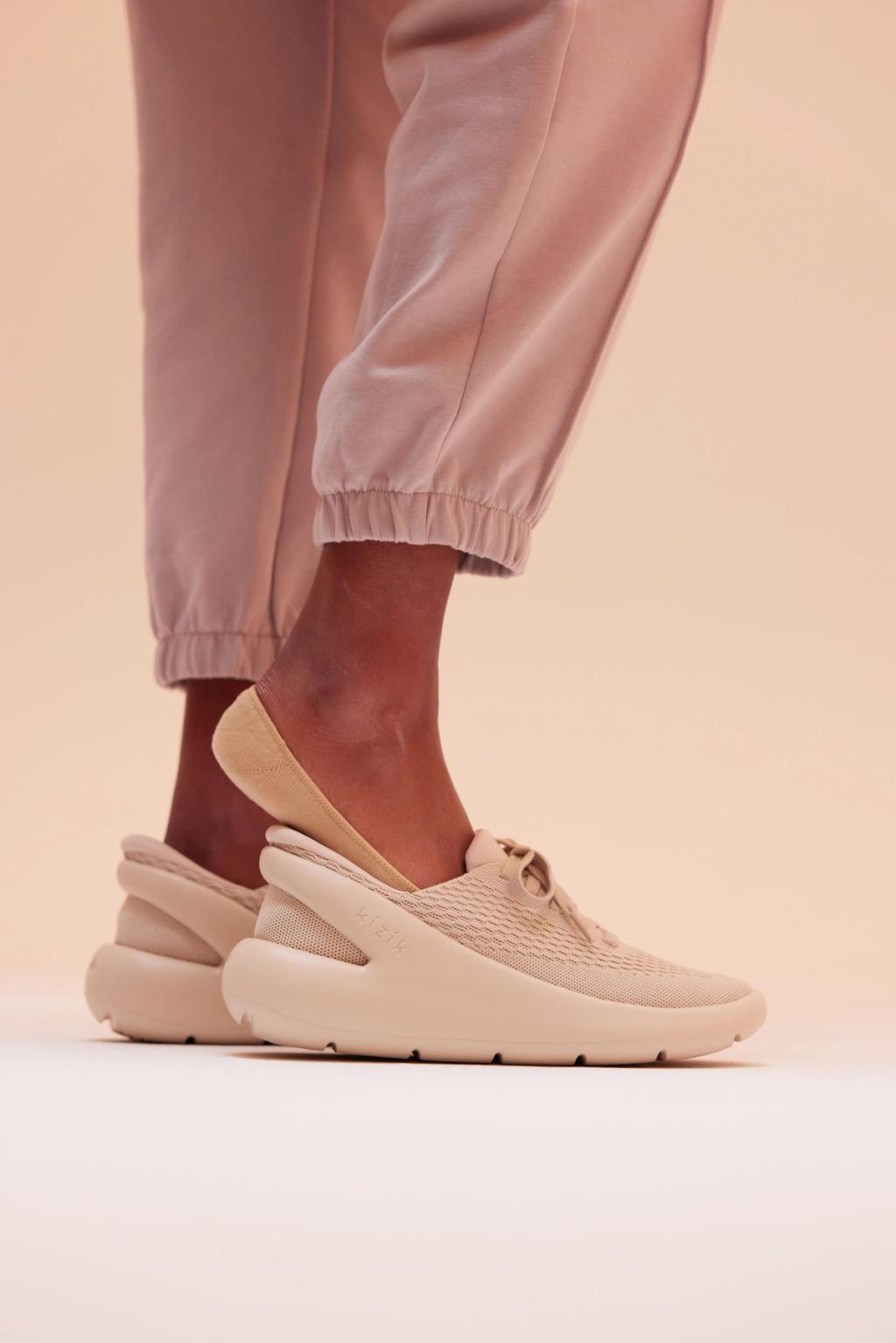 Person in pink joggers wears a tan Kizik slip-on sneaker against a neutral backdrop