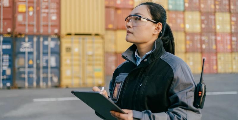 Woman in safety glasses and work jacket holds a tablet at a container yard, radio clipped to her chest.