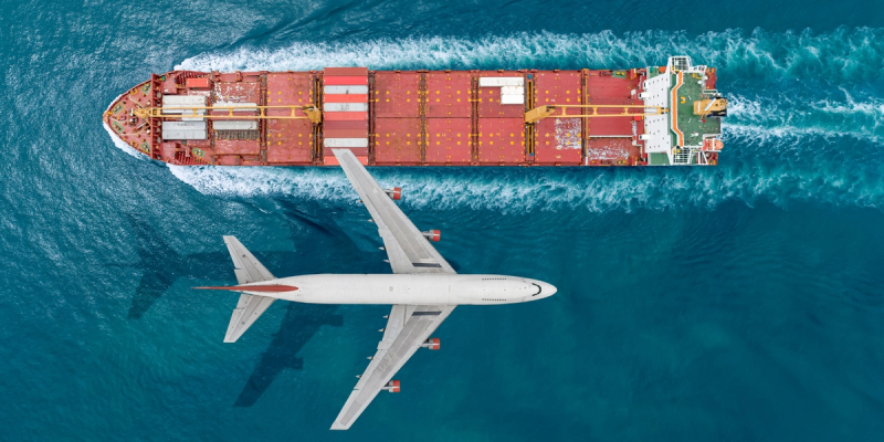 Aerial view of a red container ship at sea with a white passenger jet flying overhead