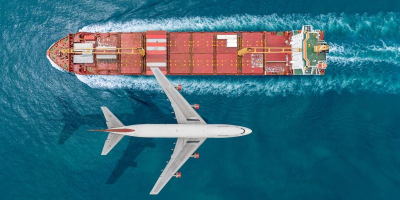 Aerial view of a red container ship at sea with a white passenger jet flying overhead