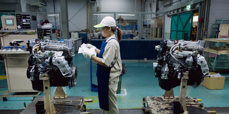 Woman in cap and apron inspecting two automotive engines mounted on stands in a manufacturing facility