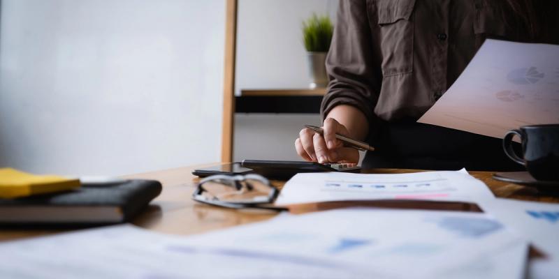 Close-up of a person at a desk using a calculator and pen with charts on paper, eyeglasses and a coffee cup nearby