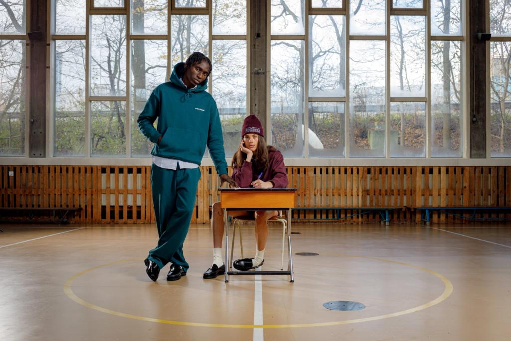 Two young models in Pockies loungewear in a gymnasium, one standing beside a seated girl at a desk