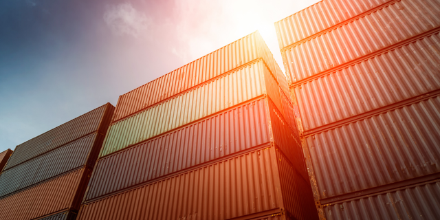 Stacks of orange shipping containers backlit by sun flare against a dusk sky, evoking supply chain growth.
