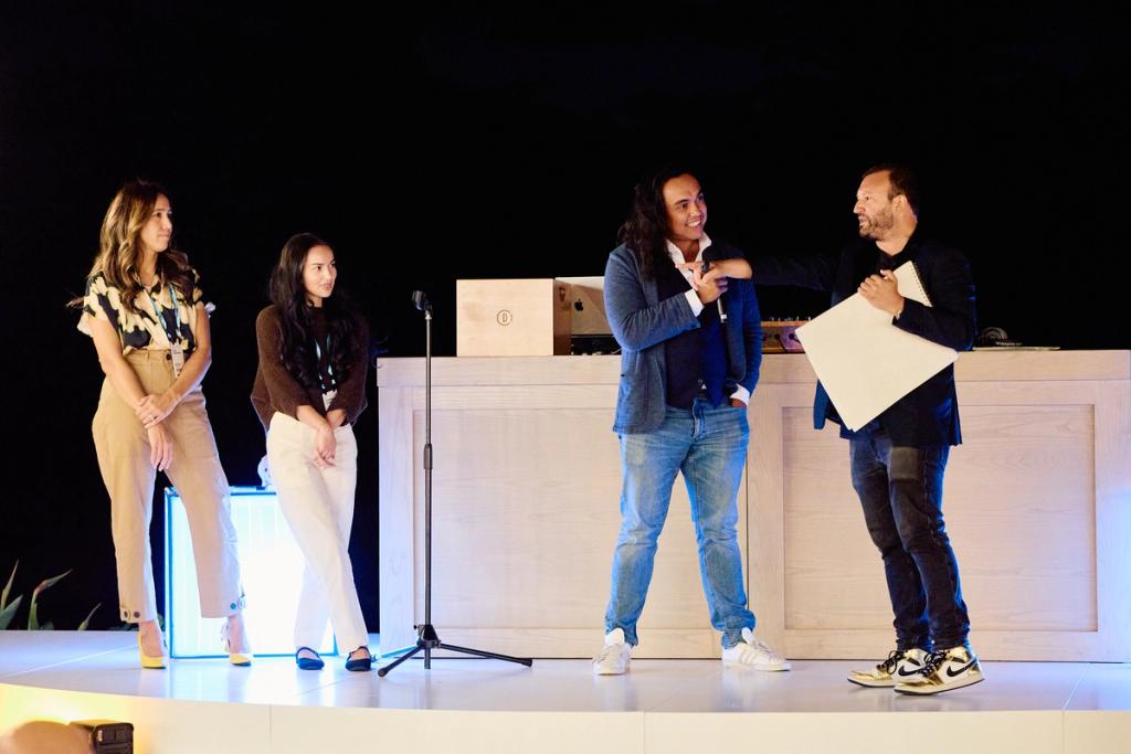 Four people on stage at an event, two reviewing a large white card under spotlights