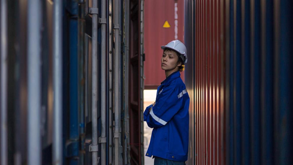 Worker in blue jacket and white hard hat with arms crossed standing in a narrow aisle between stacked shipping containers