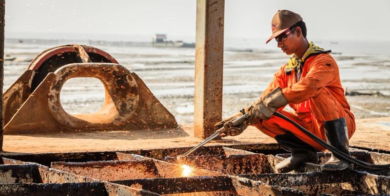 Worker in an orange suit cutting metal with a torch sending sparks at a coastal scrapyard