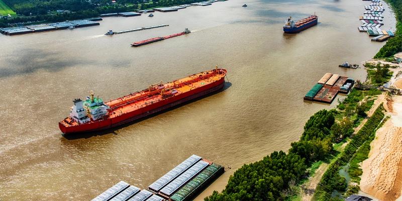 Aerial view of a red oil tanker and cargo barges on a wide brown river beside green banks