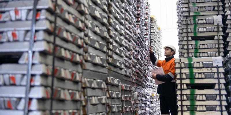Worker in safety vest and white helmet inspecting tall stacks of stored metal ingots in a warehouse