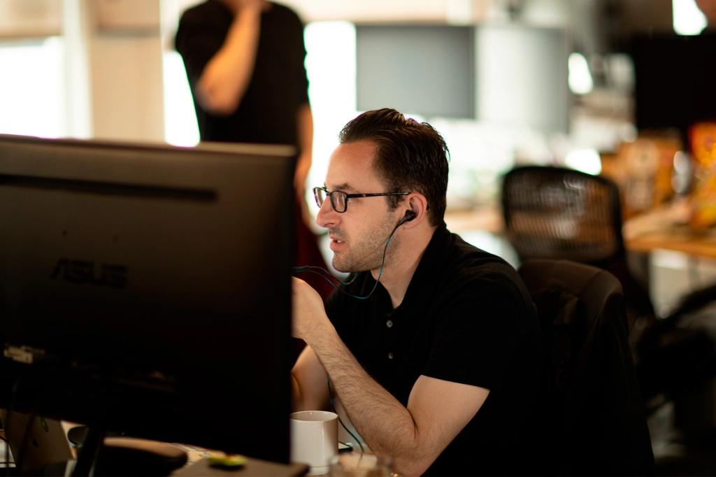 Man in glasses and earbuds works intently at an ASUS monitor in a dim open-plan office