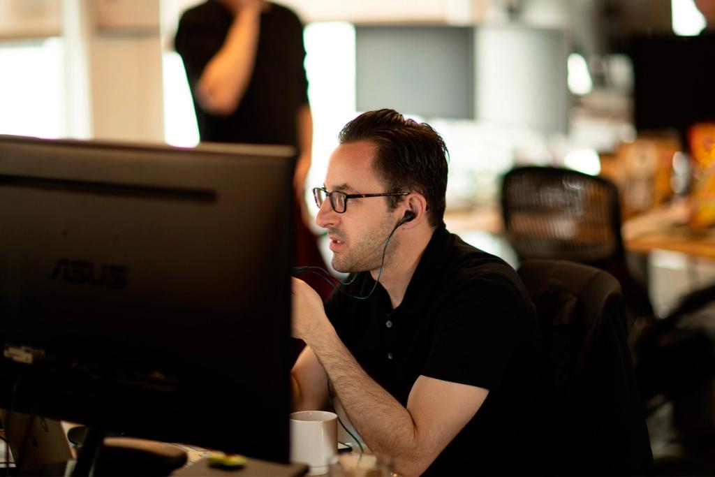 Man in glasses and earbuds works intently at an ASUS monitor in a dim open-plan office