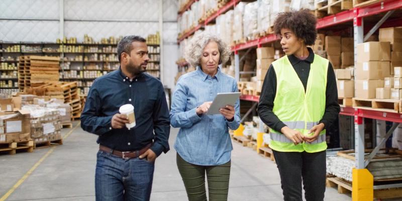 Three colleagues walk and talk in a warehouse aisle, one in a hi-vis vest, one reviewing a tablet