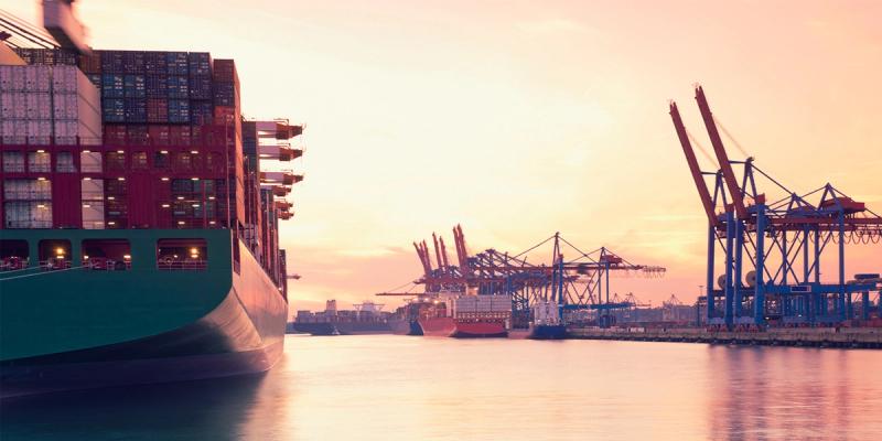 Two men in hard hats and high-visibility vests inspect stacked shipping containers in a port yard