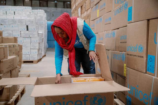 Woman in a red headscarf and UNICEF vest opening a supply box in a warehouse stacked with UNICEF cartons