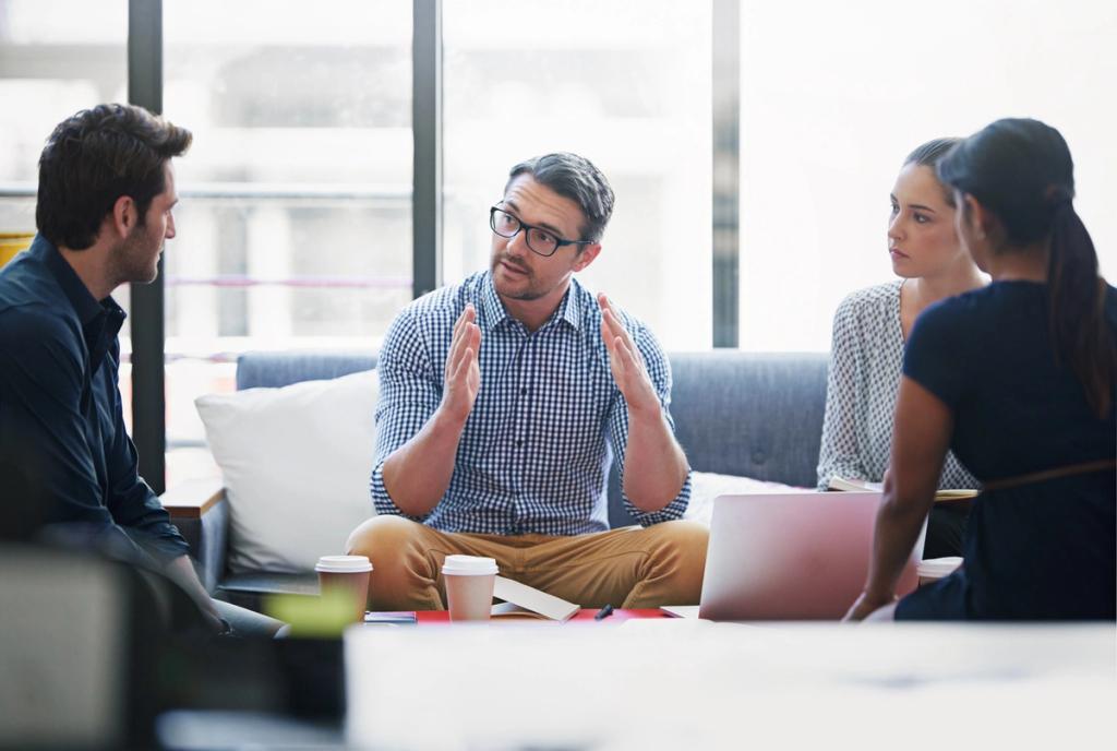 Four professionals collaborating in a meeting, with a man in a checkered shirt gesturing while speaking.