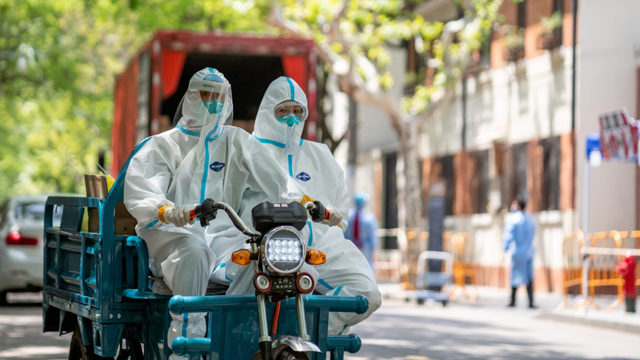 Two workers in white protective hazmat suits and masks ride a cargo motorcycle down an empty city street