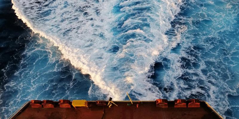 Aerial view from a ship's stern over the rust-colored deck into churning white wake on deep blue ocean